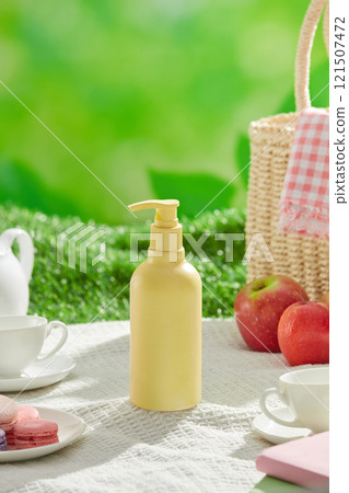 A template image with a picnic landscape theme, a white carpet with a tea set, macaroons, apples, books, baskets, and an empty label pump bottle in yellow color, on green grass. Frontal view. A template image with a picnic landscape theme, a white carpet with a tea set, macaroons, apples, books, baskets, and an empty label pump bottle in yellow color, on green grass. Frontal view. 121507472