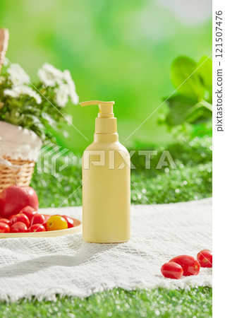 A white picnic mat is laid out in a verdant meadow, along with a daisy basket, a plate with an apple and some tomatoes, and the front of a yellow, unbranded bottle. Cosmetics advertising photo. 121507476