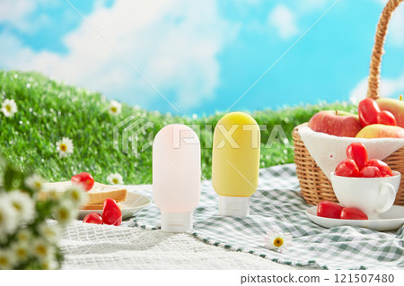 Frontal shot of two bottle mockups displayed on a blue picnic rug, next to an apple basket and some picnic utensils. A picnic concept on a green meadow with some daisy flowers in a blue sky. 121507480