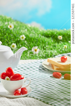 A green picnic mat with a variety of picnic items spread out on it, and empty space for displayed products. A picnic outside on a patch of green grass with daisy blooms, under a blue sky and sunshine. 121507481