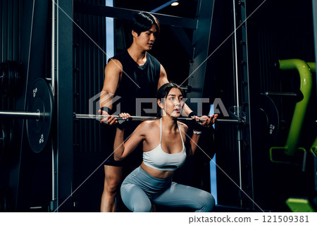 Asian woman guy a squat exercise legs heavy weights with a barbell with the assistance of a trainer in gym interior, Man doing training lifting barbell to female at fitness gym center, sport workout 121509381