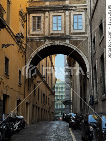 Visitors stroll down a narrow street in Rome, where a historical arch connects two buildings. Motorcycles are parked along the cobblestone road, creating a quintessential Italian atmosphere. Visitors stroll down a narrow street in Rome, where a historical arch connects two buildings. Motorcycles are parked along the cobblestone road, creating a quintessential Italian atmosphere. 121510299