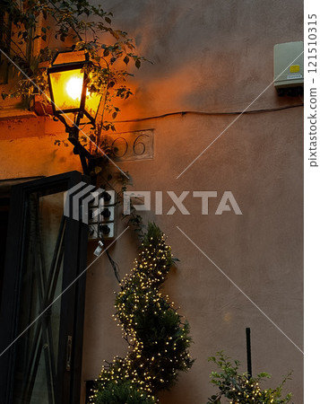 A vintage streetlamp casts a warm orange light on a charming entrance. Twinkling lights wrap around small greenery, enhancing the inviting atmosphere at dusk in a quiet neighborhood. A vintage streetlamp casts a warm orange light on a charming entrance. Twinkling lights wrap around small greenery, enhancing the inviting atmosphere at dusk in a quiet neighborhood. 121510315