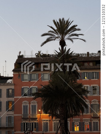 Tall palm trees create a striking silhouette as the sun sets behind colorful buildings in a coastal city. Warm lights illuminate the balconies, enhancing the tranquil evening atmosphere. Tall palm trees create a striking silhouette as the sun sets behind colorful buildings in a coastal city. Warm lights illuminate the balconies, enhancing the tranquil evening atmosphere. 121510332