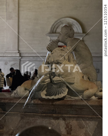 Visitors admire the detailed stone fountain adorned with figures and flowing water in a lively public square illuminated by soft lighting during the evening. 121510354