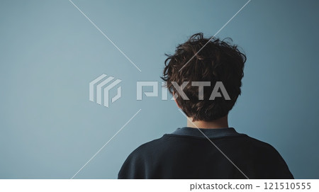 A young individual with curly hair is positioned with their back to the viewer, contemplatively gazing at a soft blue wall in a well-lit indoor space, suggesting reflection or deep thought. 121510555
