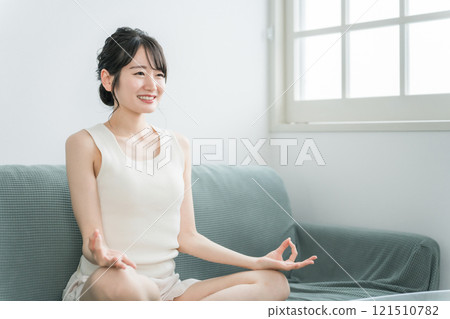 年輕的亞洲女性在家裡的沙發上做輕瑜伽、冥想、正念、晨間活動（晨間活動） 121510782