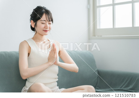 年輕的亞洲女性在家裡的沙發上做輕瑜伽、冥想、正念、晨間活動（晨間活動） 121510783