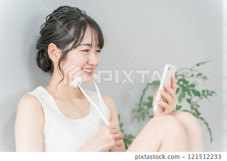 A smiling Asian woman relaxes looking at her smartphone while massaging her face with a facial roller at home 121512233