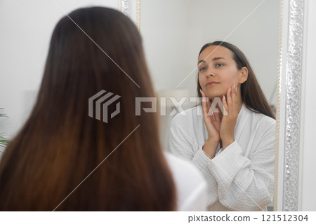 A woman, in her cozy bathrobe, practices a beauty skincare routine at home, studying herself in the bathroom mirror. A woman, in her cozy bathrobe, practices a beauty skincare routine at home, studying herself in the bathroom mirror. 121512304