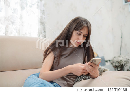 Young Caucasian woman sitting comfortably in a chair, smiling while using her smartphone indoors during a relaxing afternoon 121516486