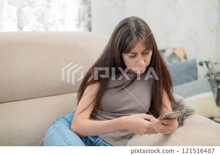 Young woman sitting comfortably in a chair, smiling while using her smartphone indoors during a relaxing afternoon 121516487