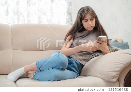 Young Caucasian woman sitting comfortably in a chair, smiling while using her smartphone indoors during a relaxing afternoon 121516488
