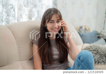 Young Caucasian woman sitting comfortably on sofa, talking with smartphone indoors during a relaxing afternoon 121516505