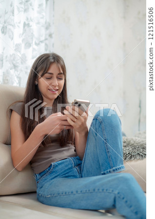 Young happy woman sitting comfortably in a chair, smiling while using her phone indoors during a relaxing afternoon 121516506
