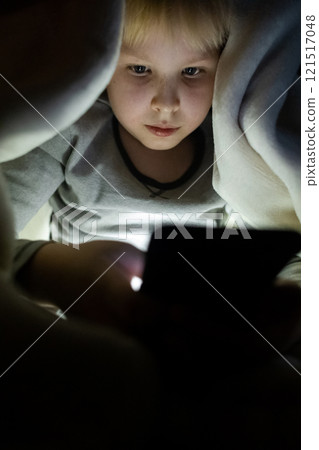 Child phone addiction. The boy at night, covered with a blanket, on the bed plays games on the smartphone and watches videos, cartoons 121517048