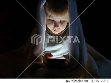 Child phone addiction. The boy at night, covered with a blanket, on the bed plays games on the smartphone and watches videos, cartoons Child phone addiction. The boy at night, covered with a blanket, on the bed plays games on the smartphone and watches videos, cartoons 121517055