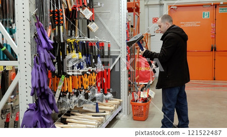 Customer selecting sledgehammer, testing weight, comparing sizes near shopping basket amid hardware store displays of construction tools and professional equipment. Translation: Sale 121522487