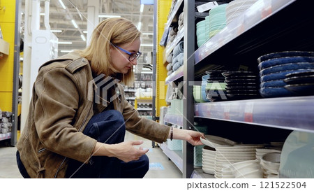 Woman selecting plates in a supermarket houseware section, holding a white plate and browsing the shelves 121522504