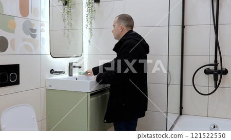 Man washing hands in modern bathroom, green vanity unit, illuminated mirror, black fixtures highlighting contemporary design aesthetic Man washing hands in modern bathroom, green vanity unit, illuminated mirror, black fixtures highlighting contemporary design aesthetic 121522550