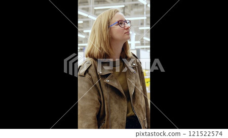 Young woman with glasses and a brown leather jacket gazes upwards while browsing in a warehouse store with fluorescent lighting 121522574