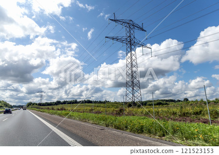 High voltage power line pylons, electrical tower on a green field with blue sky. Highway with cars on a cloudy summer day. High voltage power line pylons, electrical tower on a green field with blue sky. Highway with cars on a cloudy summer day. 121523153