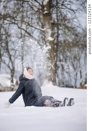 Young woman sits in the snow, tossing a mound of snow over her head in a winter woodland 121524134