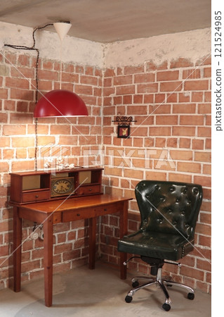 Dark vintage chair on wheels against brick wall next to wooden table on which there is shelf with old clock over which large red lamp hangs Dark vintage chair on wheels against brick wall next to wooden table on which there is shelf with old clock over which large red lamp hangs 121524985