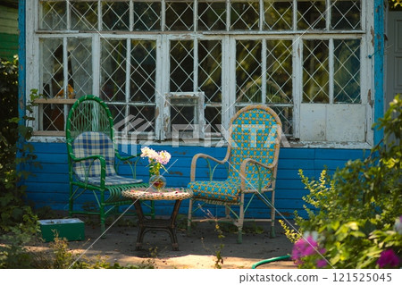 Wicker chairs against background of old country house near round table on which there is glass vase with white flowers inside. 121525045