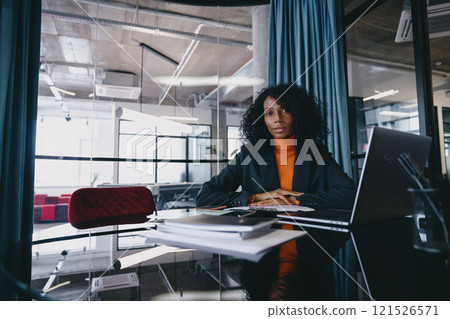 Serious Black businesswoman, wearing stylish blazer and vibrant orange sweater, sits at office desk filled with documents, contemplating while looking at the camera, in a well-lit modern workspace. Serious Black businesswoman, wearing stylish blazer and vibrant orange sweater, sits at office desk filled with documents, contemplating while looking at the camera, in a well-lit modern workspace. 121526571
