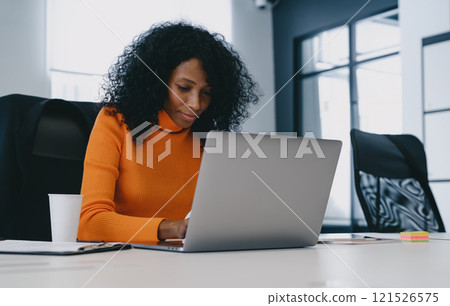Concentrated professional woman in a bright orange top engages with laptop in a modern office, embodying focused productivity. Concentrated professional woman in a bright orange top engages with laptop in a modern office, embodying focused productivity. 121526575