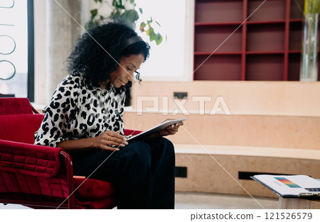 Black businesswoman in leopard print blouse focused on tablet in a modern office lounge, merging style with digital proficiency in a relaxed setting. 121526579