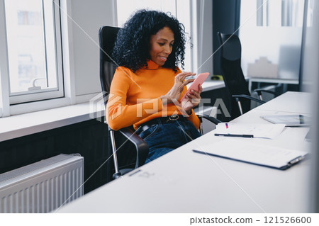 Smiling Black businesswoman in her 30s, enjoying a light moment with her smartphone in a sunny, modern office, reflecting a relaxed work environment in autumn. 121526600