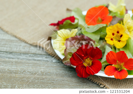 Salad of multicolored edible nasturtium flowers and lettuce in a plate on the burlap mat on the wooden table. 121531204