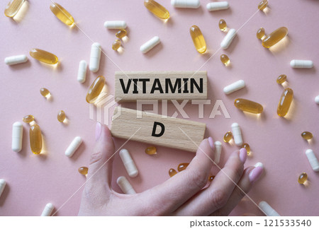 Vitamin D symbol. Concept word Vitamin D on wooden blocks. Beautiful pink background with pills. Doctor hand. Medicine and Vitamin D concept. Copy space Vitamin D symbol. Concept word Vitamin D on wooden blocks. Beautiful pink background with pills. Doctor hand. Medicine and Vitamin D concept. Copy space 121533540