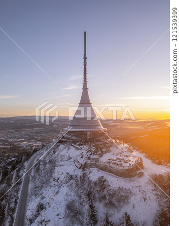 A breathtaking winter sunrise illuminates Jested Mountain, highlighting its unique architectural structure against a backdrop of snowy terrain and a clear sky. 121539399
