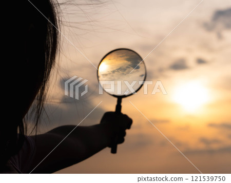Cute little girl holding magnifying glass in garden on sunset sky background. 121539750