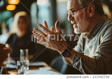 A man is talking to someone at a table. He is wearing glasses and has a watch on his wrist. A man is talking to someone at a table. He is wearing glasses and has a watch on his wrist. 121542730
