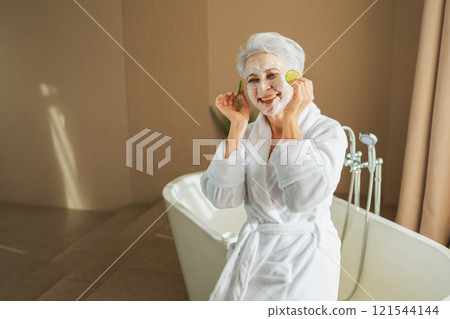 Mature adult old woman with nourishing anti age face cream mask holding cucumber slices. Senior woman with white mask creme on skin gets facial treatment daily beauty routine. Skin care aging beauty Mature adult old woman with nourishing anti age face cream mask holding cucumber slices. Senior woman with white mask creme on skin gets facial treatment daily beauty routine. Skin care aging beauty 121544144