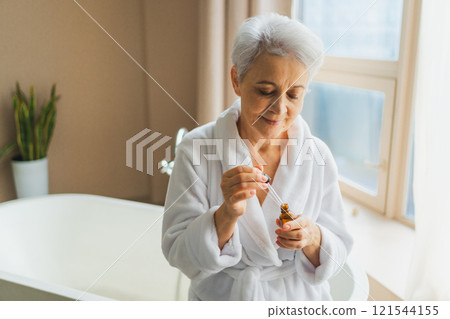 Woman applying natural drops cosmetics oil to face skin. Mature adult old woman applying nourishing anti age serum with pipette doing daily beauty routine. Senior woman gets kin scare facial treatment 121544155