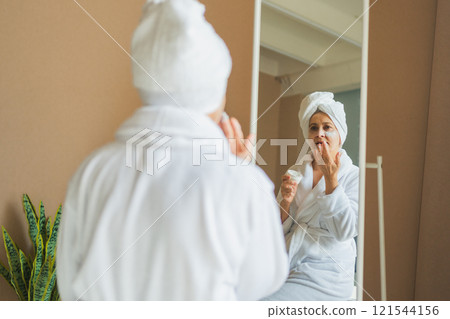Mature adult old woman looking at mirror applying nourishing anti age face cream doing daily beauty routine. Senior woman with white mask creme on skin gets facial treatment Skin care aging skincare Mature adult old woman looking at mirror applying nourishing anti age face cream doing daily beauty routine. Senior woman with white mask creme on skin gets facial treatment Skin care aging skincare 121544156