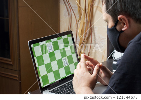 A man in a medical mask solves a chess problem online. Chess board with figures on a laptop screen. Leisure in self-isolation. International chess day. A man in a medical mask solves a chess problem online. Chess board with figures on a laptop screen. Leisure in self-isolation. International chess day. 121544675