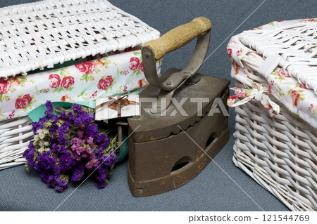 Old iron, heated by hot coals. Located on gray fabric. Nearby are wicker baskets, a bouquet of dried flowers and a greeting card. Old iron, heated by hot coals. Located on gray fabric. Nearby are wicker baskets, a bouquet of dried flowers and a greeting card. 121544769