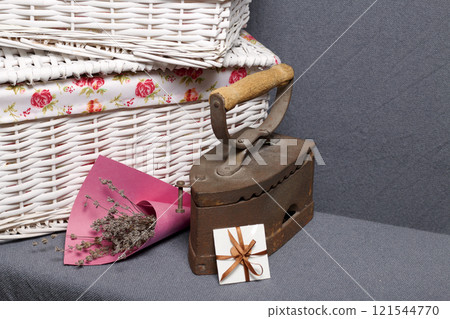 Old iron, heated by hot coals. Located on gray fabric. Nearby are wicker baskets, a bouquet of dried flowers and a greeting card. Old iron, heated by hot coals. Located on gray fabric. Nearby are wicker baskets, a bouquet of dried flowers and a greeting card. 121544770