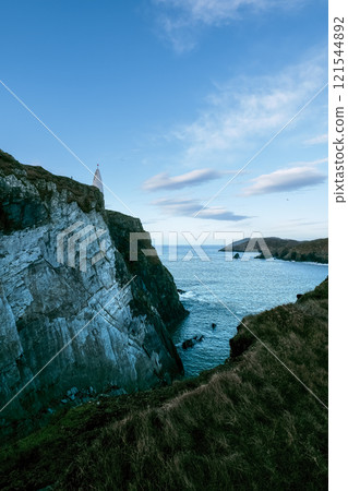 View of the lighthouse on a rocky coastal cliff by the sea under a cloudy sky at dusk. View of the lighthouse on a rocky coastal cliff by the sea under a cloudy sky at dusk. 121544892