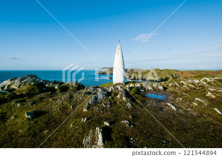 Ocean view. A tall, slender, white cone-shaped tower stands prominently on a rocky hill overlooking a tranquil ocean. Ocean view. A tall, slender, white cone-shaped tower stands prominently on a rocky hill overlooking a tranquil ocean. 121544894