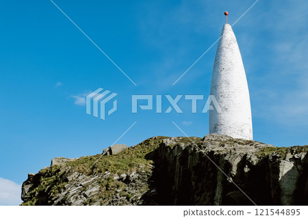 This photo depicts a tall, white, cylindrical structure situated on a cliff top overlooking a clear blue expanse of sky. This photo depicts a tall, white, cylindrical structure situated on a cliff top overlooking a clear blue expanse of sky. 121544895