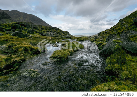 Rocky terrain surrounds a flowing stream. Vegetation thrives along the water's edge. A distant view is visible. Rocky terrain surrounds a flowing stream. Vegetation thrives along the water's edge. A distant view is visible. 121544896
