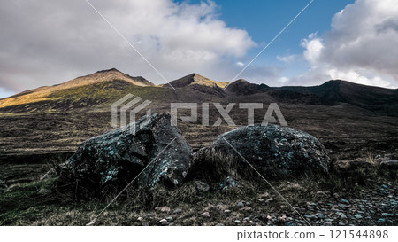 Foreground displays two dark, rounded rocks amidst sparse vegetation. Background shows a mountainous terrain under a blue sky with clouds. 121544898