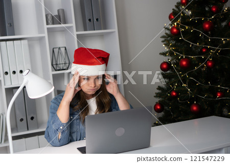 During the holidays, a frustrated person wears a Santa hat while sitting at their laptop, trying to balance work and Christmas joys, struggling with motivation and technology challenges 121547229